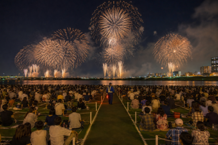 Spectators sit in managed riverbank sections beside a clear emergency aisle while professional Japanese fireworks fill the night sky across the river.