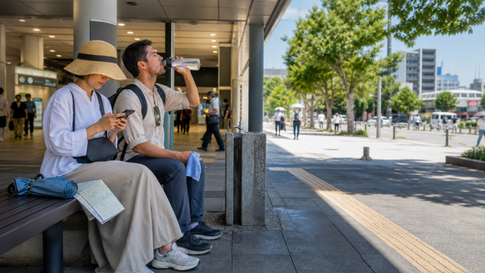 Two adult travelers rest in the shade of a Japanese station, drink water and check their route before entering the midsummer sun