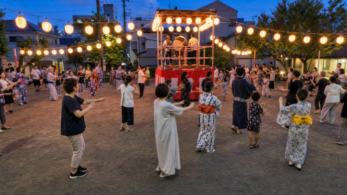 Adults and families in yukata and everyday summer clothes dance in an orderly circle around a neighborhood bon odori yagura at blue hour