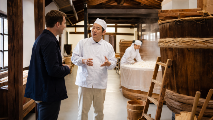 Adult traveler learning about steamed rice and cedar vats from a guide inside a traditional Japanese sake brewery
