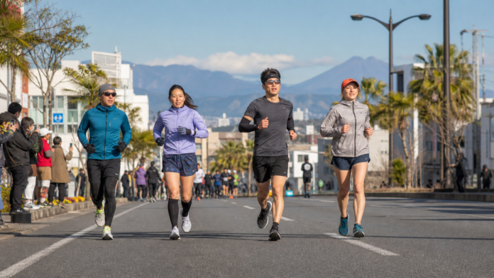 Four recreational runners compete on a southern Japanese city road in cool winter sunlight, with spectators, subtropical street trees and volcanic hills