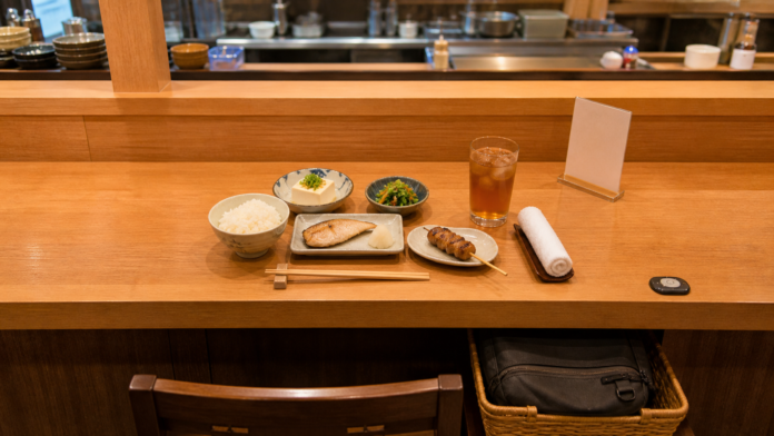 One solo place setting at a Japanese izakaya counter with small dishes, oolong tea, call button and a travel bag stored in a basket